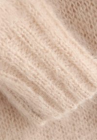 Tessuto lavorato a maglia beige chiaro con una texture morbida e pelosa. Presenta motivi a coste e un design a trama sciolta per garantire calore.