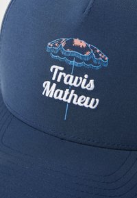 Navy cap featuring white and blue embroidered text "Travis Mathew" beneath a colorful umbrella design. Smooth fabric with structured shape.