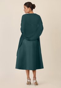 Türkises Satin-Kleid, knielang, mit fließender Silhouette und zarter Drapierung. Ein transparenter Schal, der über die Schultern drapiert ist, kombiniert mit silbernen, rückenfreien High Heels.
