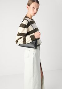 Svart triangulär läderväska hålls av en modell som bär en krämfärgad och mörkgrön randig tröja samt en ljus denimkjol med en sidoslits.