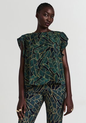Donna che indossa un completo nero coordinato con top e pantaloni decorati da motivi di foglie verdi e gialle, in piedi davanti a uno sfondo chiaro e uniforme.
