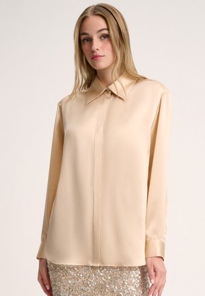Beige silkipuseri, jossa on terävä kaulus, etupainatuskiinnitys ja pitkät hihat. Sujuva tekstuuri, ei kuvioita, yhdistettynä paljettihameeseen.