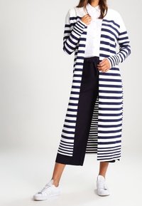 Žena ve dlouhém cardiganovém svetru s námořnickými a bílými pruhy, bílou košili na knoflíky, námořnickými culottes a bílými teniskami, stojící na jednobarevném pozadí.