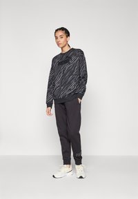 Pullover nero e grigio con un motivo ondulato, caratterizzato da un logo sul petto, abbinato a pantaloni scuri e sneaker chiare.