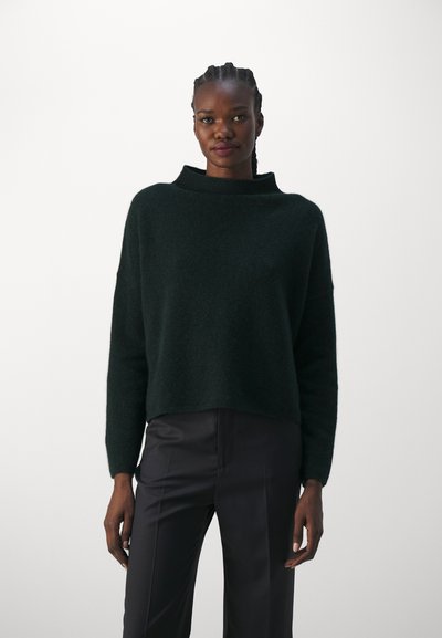 Maglione corto verde scuro realizzato in morbido materiale testurizzato, con scollatura alta e maniche lunghe. Abbinato a pantaloni neri lisci.