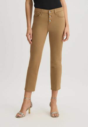 Jeans skinny marron taille haute, fermeture à cinq boutons et poches avant. Portés avec des sandales à talons hauts argentées et à lanières.