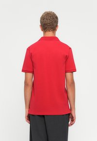 Mann mit lockigem Haar steht mit dem Rücken zur Kamera, trägt ein rotes kurzärmeliges Poloshirt und dunkle graue Hosen vor einem schlichten hellen Hintergrund.