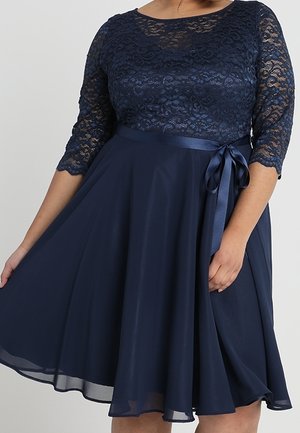 Žena oblečená v námořnicky modrých šatech po kolena s krajkovým živůtkem, tříčtvrtečními rukávy a saténovou stuhou uvázanou v pase.