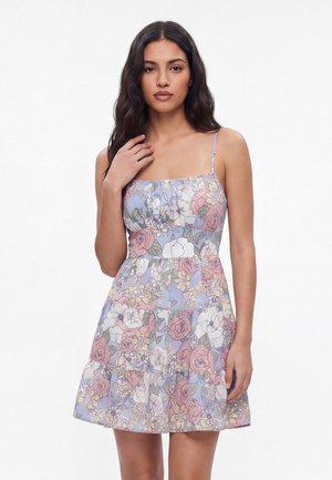 Kvinde med langt mørkt hår iført en ærmeløs pastelblomstret kjole med tynde stropper, stående mod en hvid baggrund.