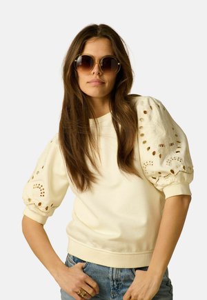 Femme aux longs cheveux bruns porte de grandes lunettes de soleil rondes et un haut crème à manches bouffantes avec broderie ajourée, assorti à un jean bleu.