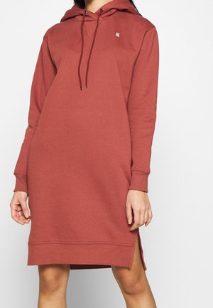 Femme portant une robe sweat à capuche ample manches longues de couleur brun rougeâtre avec des fentes latérales et un petit logo sur la poitrine.