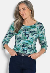 Langärmliges floral bedrucktes Oberteil in Grün-, Blau- und Rosatönen. Mit einem Rundhalsausschnitt und einem lässigen Schnitt, kombiniert mit dunkelblauen Hosen und einem Gürtel.