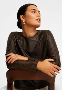 Bronzefärgad, texturerad långärmad topp med rund halsringning. Modellen sitter med armarna i kors på en trästol och bär guldhoops.
