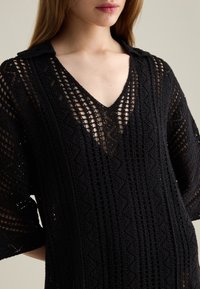 Schwarzes Häkelkleid mit V-Ausschnitt, lockeren Ärmeln, strukturiertem Muster und offenem Strickdesign für Atmungsaktivität und Stil.