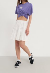Jeune femme portant un crop top violet noué, une jupe blanche à volants et des baskets Adidas noir et blanc, se tenant devant un fond uni.