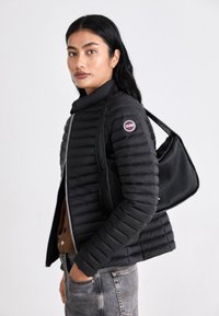 Melnās pūkainās jakas ar horizontālu quilted dizainu, augsto apkakli un priekšējo rāvējslēdzēju. Apvienota ar teksturētu melnu plecu soma.