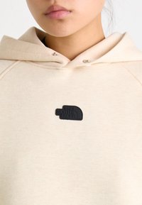 Bežs kapucis, kas izgatavots no mīksta materiāla ar melnu gumijas logo uzlīmi krūšu daļā. Tam ir kapuce un sudraba spiedpoges detaļas.