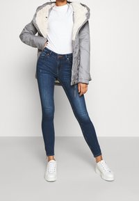 Grå isolerad jacka med en mjuk, vit pälsfoder, kombinerad med välsittande mörkblå jeans och vita sneakers. Dragkedjedetaljer på jeansen.