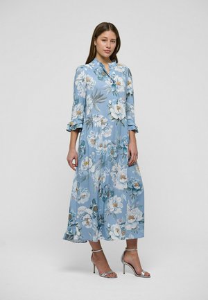 Donna in piedi che indossa un abito midi azzurro a fiori con polsini arricciati e sandali argentati con tacco alto su uno sfondo neutro.