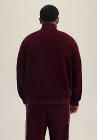 Burgundowa kurtka zapinana na zamek z teksturowanym wzorem, prążkowanymi mankietami i kołnierzem, z bocznymi paskami i małym logo z tyłu.