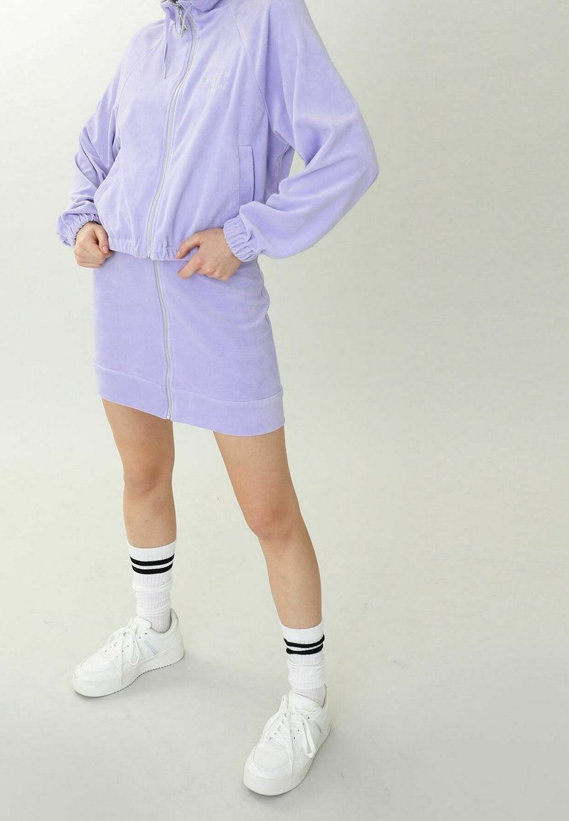 Lila velour zip-up jacka med matchande mini kjol. Outfiten har en avslappnad passform och elastiska ärmslut. Bärs med vita sneakers och randiga strumpor.