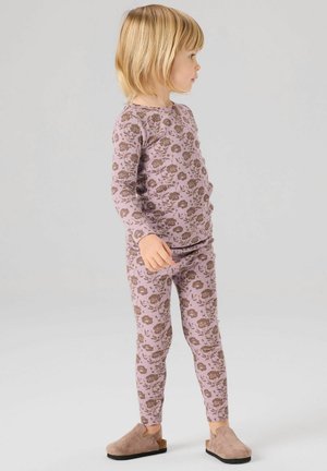 Kind mit blondem Haar, das ein langärmliges, lila Oberteil mit Blumenmuster und passende Leggings sowie beige Slipper trägt, steht seitlich.