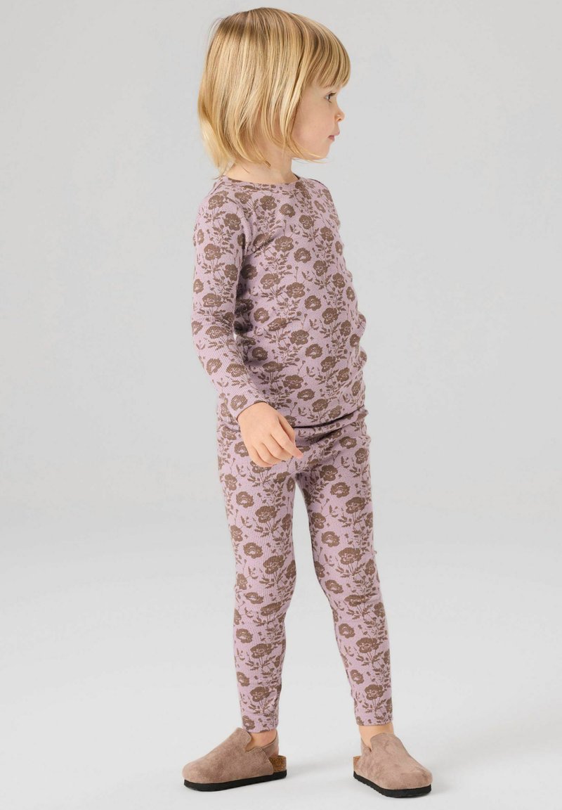 Enfant aux cheveux blonds portant un haut à manches longues violet à motifs floraux et un legging assorti, avec des chaussures beige à enfiler, debout de côté.