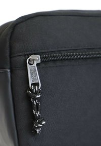 Clip de mochila de tela negra con cierre de cremallera, que presenta una pestaña plateada marcada con "coronel tapioca" y un cordón trenzado en negro y blanco.