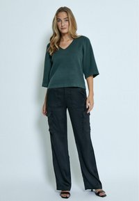 Maglione verde scuro a maniche tre quarti con scollo a V abbinato a pantaloni cargo neri a gamba larga dotati di tasche laterali e una texture liscia.