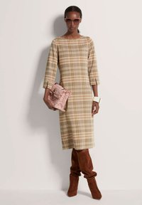 Langmouwige, geruite jurk in beige en bruine tinten, gecombineerd met een roze gestructeerde clutch en hoge bruine suède laarzen.