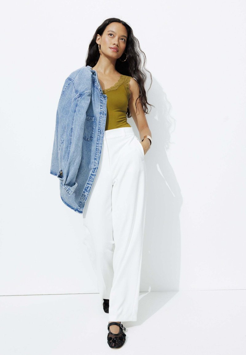 Débardeur vert olive avec dentelle, pantalons blancs taille haute, veste en denim délavée portée sur l'épaule, et chaussures noires ornées.