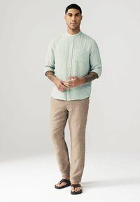 Camisa de manga larga texturizada verde claro con cuello mao, combinada con pantalones de lino beige y chanclas negras. Diseño sencillo, ajuste casual.