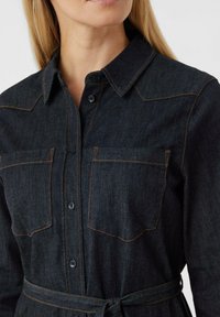 Ciemnoniebieska koszula dżinsowa z kołnierzykiem, dwoma przednimi kieszeniami i pomarańczowymi przeszyciami. Posiada paskowany w pasie krój, zapewniający dopasowanie.