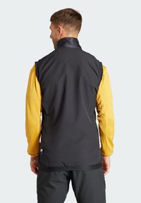 Schwarze ärmellose Weste mit einem strukturierten Kragen, getragen über einem gelben Langarmshirt. Verfügt über einen glatten Stoff und ein Logo-Patch an der Seite.