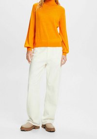 Pull orange à manches longues avec un col montant, associé à un pantalon blanc à jambes larges et des chaussures beige sans lacets, présentant un tissu texturé.