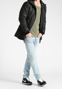 Man i svart vadderad jacka, grön stickad tröja, ljusblå jeans och svart-vita Nike-sneakers, står med händerna i fickorna.