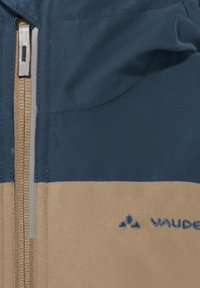 Veste zippée bleu marine et beige avec un design bicolore, un tissu texturé et un logo sur le côté inférieur gauche. Matériau lisse et résistant à l'eau.