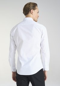 Biała koszula z długim rękawem, wzorzysta w czarne kropki, z kołnierzykiem zapinanym na guziki i zaokrąglonym dołem. Materiał wydaje się gładki i lekki.