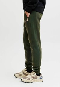 Grønne sweatpants med ribbede sidepaneler, elastisk talje og lynlåslommer. Båret med beige og sorte sneakers.