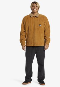 Brun corduroy jakke med en tan faux fur krave, frontlomme og en afslappet pasform, parret med mørke bukser og sorte sneakers.