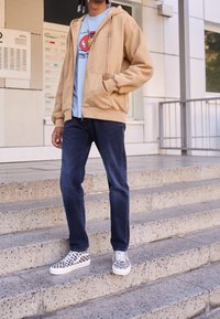 Sudadera beige con cremallera sobre una camiseta gráfica azul clara, jeans azules y zapatillas de cuadros en blanco y negro, de pie en unos escalones exteriores.