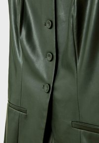 Giacca in pelle verde con una texture liscia, dotata di grandi bottoni, due tasche laterali e un design su misura che mostra linee pulite.