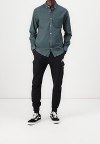 Uomo che indossa una camicia a bottoni verde scuro, pantaloni cargo neri con tasche e scarpe da ginnastica nere e bianche, in piedi davanti a uno sfondo bianco uniforme.