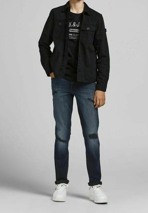 Persona con giacca nera con bottoni, t-shirt grafica nera, jeans blu scuro con chiazze sbiadite e sneakers bianche, in piedi contro uno sfondo grigio.