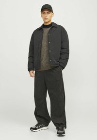 Doudoune noire avec boutons-pression sur un pull en laine brun texturé, associée à un pantalon noir ample et des baskets avec finition en mesh.
