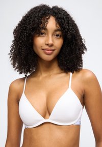 Γυναίκα με σγουρά μαλλιά που φοράει λευκό σουτιέν τριγωνάκι, χαμογελώντας απαλά, με φόντο απλό και φωτεινό.