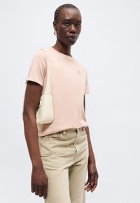 Μαύρη γυναίκα που φοράει ένα ανοιχτό ροζ κοντομάνικο μπλουζάκι, μπεζ παντελόνι, χρυσούς κρίκους για σκουλαρίκια και κουβαλά μια μικρή λευκή τσάντα ώμου.