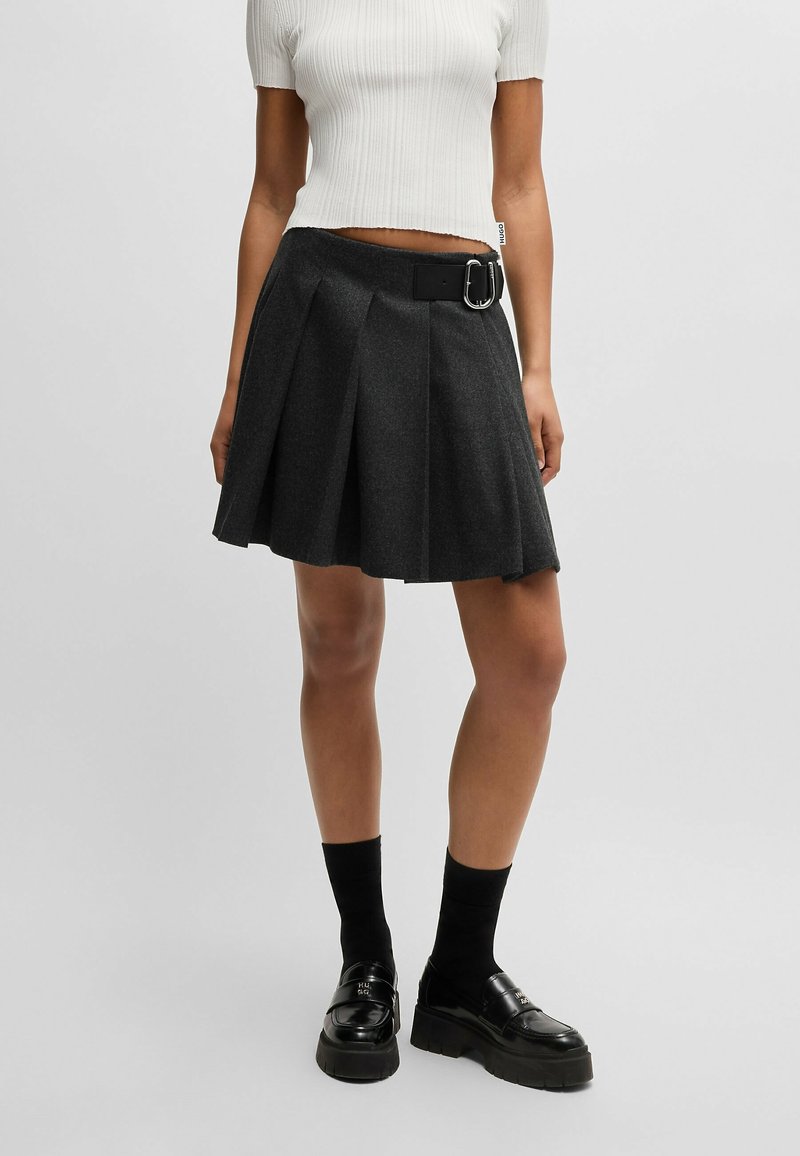 Femme portant un haut blanc à manches courtes, une jupe noire plissée avec ceinture, des chaussettes noires et des mocassins plateformes noirs sur fond gris.