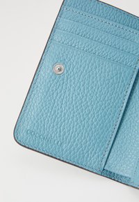 Portafoglio in pelle color teal con superficie testurizzata, quattro slot per carte, un pulsante a scatto argentato e logo impresso nell'angolo.