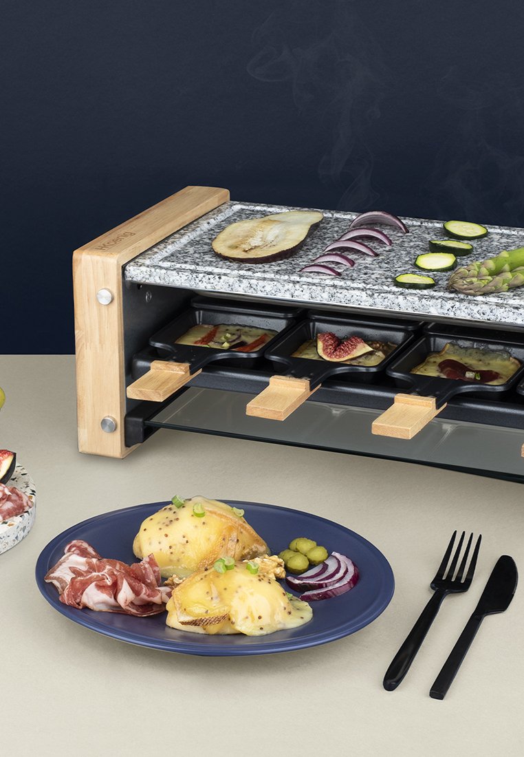 Gril à raclette avec plateau en granit et accents en bois. Comprend quatre petites plaques de cuisson. Les assiettes présentent du fromage fondu, des viandes tranchées et des légumes.